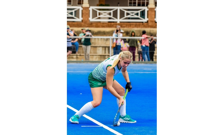 A female field hockey player in green and white uniform focuses intently as she prepares to strike a ball on a bright blue field. Spectators watch from behind a fence, adding a sense of competition.