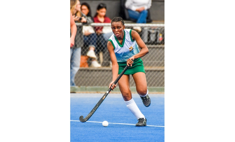 A field hockey player in a green and white uniform focuses intensely while dribbling the ball on a blue turf. Spectators sit behind a fence.