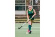 A female field hockey player in a green and yellow uniform focuses intently while pushing a ball on a turf field. She wears pink shoes.