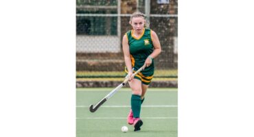 A female field hockey player in a green and yellow uniform focuses intently while pushing a ball on a turf field. She wears pink shoes.