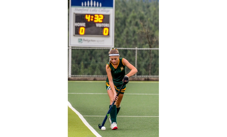 A female field hockey player in green and yellow uniform dribbles a ball on a green turf field. A scoreboard in the background shows 4:32, score 0-0.