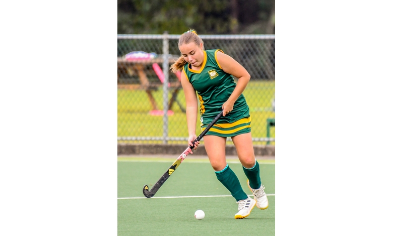 A female field hockey player in green and yellow uniform skillfully dribbles a ball on a turf field. Her focused expression highlights determination.