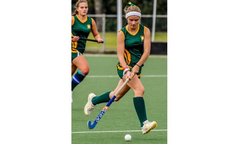 Female field hockey player in green and yellow uniform dribbling the ball with focus and agility on a grassy field, teammate in background.