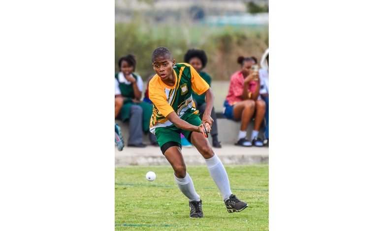 A young athlete wearing a green and yellow sports uniform intensely focuses on a ball mid-action on a grassy field, with spectators watching in the background.