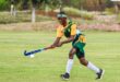 A field hockey player in green and yellow uniform skillfully hits a ball with a blue stick on a grassy field, displaying focus and athleticism.