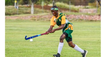 A field hockey player in green and yellow uniform skillfully hits a ball with a blue stick on a grassy field, displaying focus and athleticism.