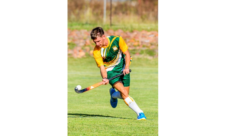 A field hockey player in a green and yellow uniform swings a stick to hit the ball. The background shows a grassy field under sunny skies.