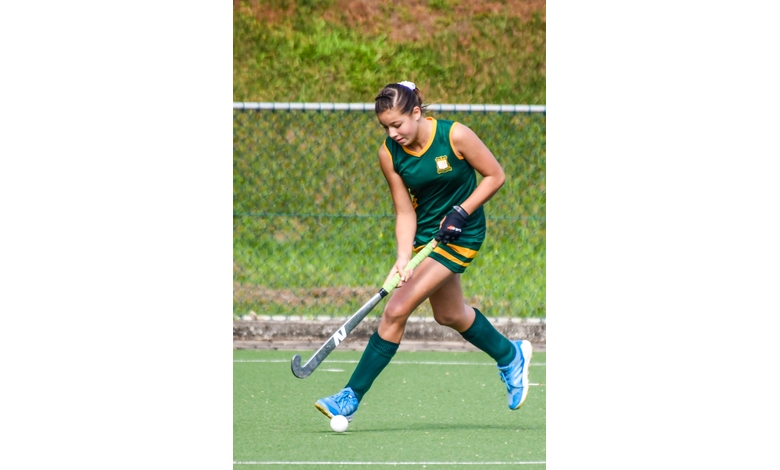 A field hockey player in a green and yellow uniform focuses intently as she skillfully maneuvers a ball with her stick on a green field.