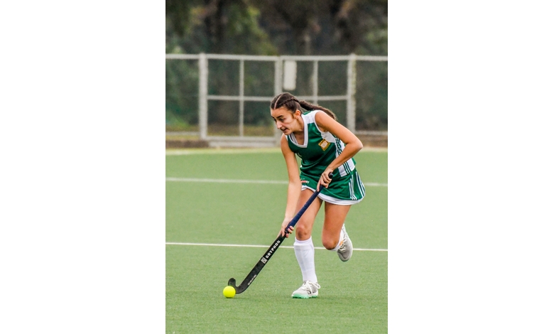 A female field hockey player in a green uniform moves across the field, focused on controlling the ball with her stick. She exudes determination and athleticism.