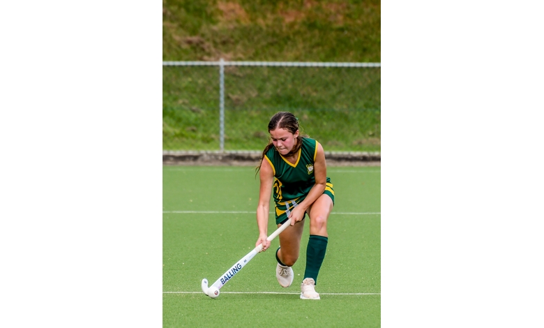 A female field hockey player in a green and yellow uniform is focused, bending slightly as she moves the ball with her stick on a grassy field, conveying determination.