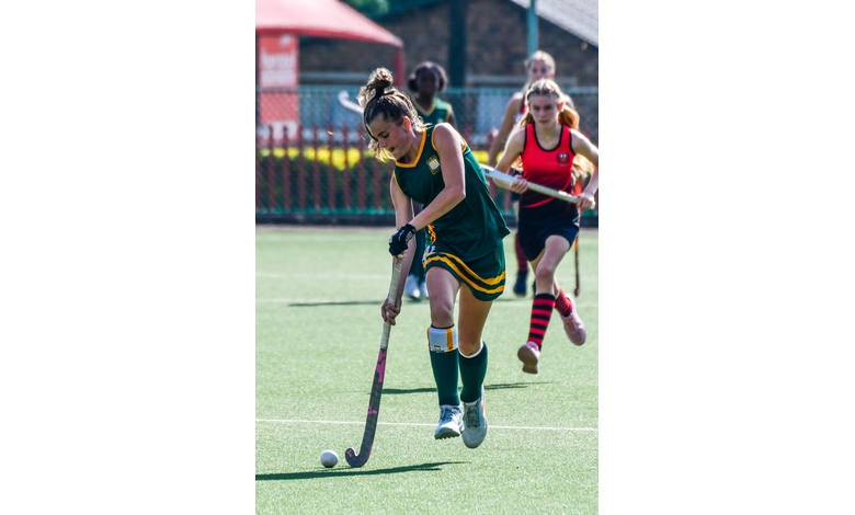 A field hockey player in a green uniform skillfully dribbles the ball. Teammates and opponents, wearing red uniforms, actively follow behind her.