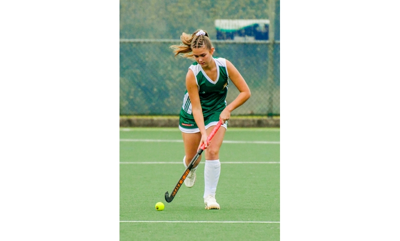 A female field hockey player in a green and white uniform is focused on dribbling a yellow ball on a green pitch, conveying concentration and athleticism.