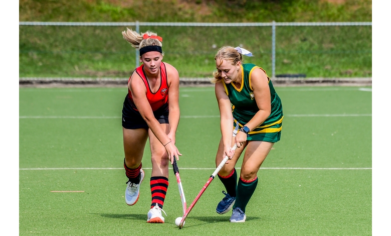Two female field hockey players in action on a grass field. One wears a red and black uniform, the other green and yellow. They focus intensely on the ball.