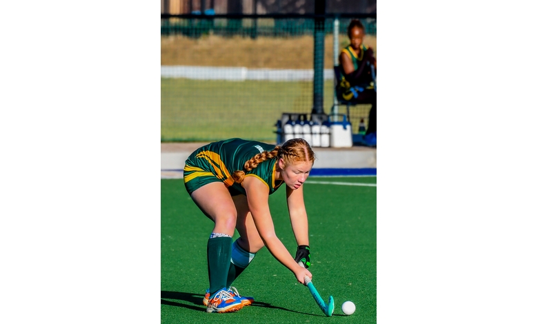 A field hockey player in a green and yellow uniform leans forward with her stick poised to hit the ball. The scene is dynamic and focused, with another player sitting in the background.