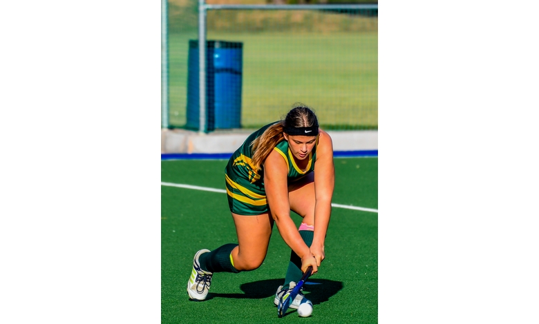 A focused field hockey player in a green and yellow uniform crouches low while handling the stick, aiming at the ball on a sunny field.