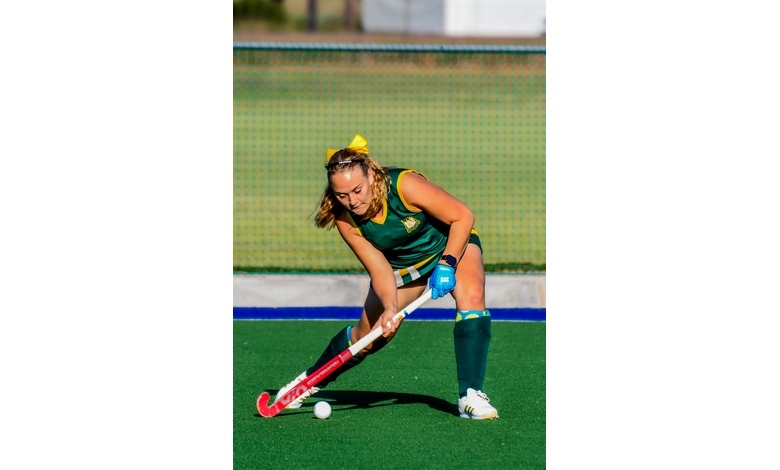 A female field hockey player in green and yellow uniform focuses intently while dribbling the ball on a bright green field. The scene conveys action and concentration.
