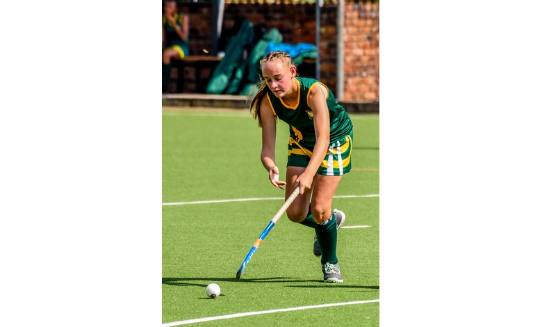 A female field hockey player in green and yellow uniform skillfully maneuvers the ball with her stick on a sunny outdoor field, displaying focus and determination.