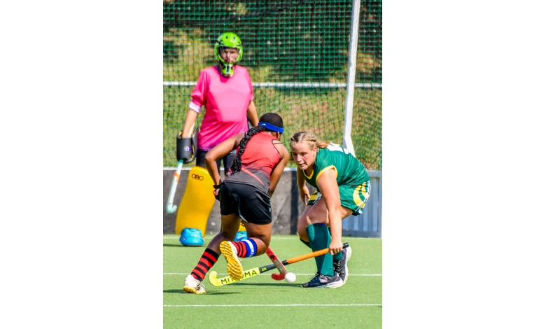 Two field hockey players in action, one in green and yellow, the other in red, compete for the ball on a green field. A goalie in bright pink observes.