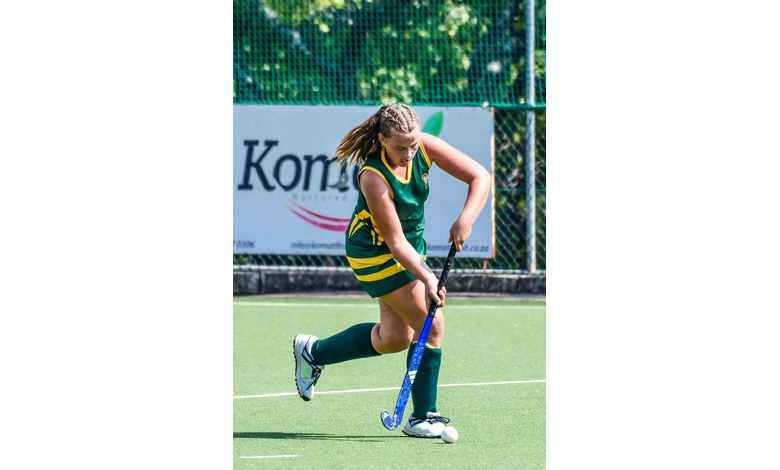 A field hockey player in green and yellow strikes the ball with focus on an outdoor turf. A blurred ad banner is visible in the background.