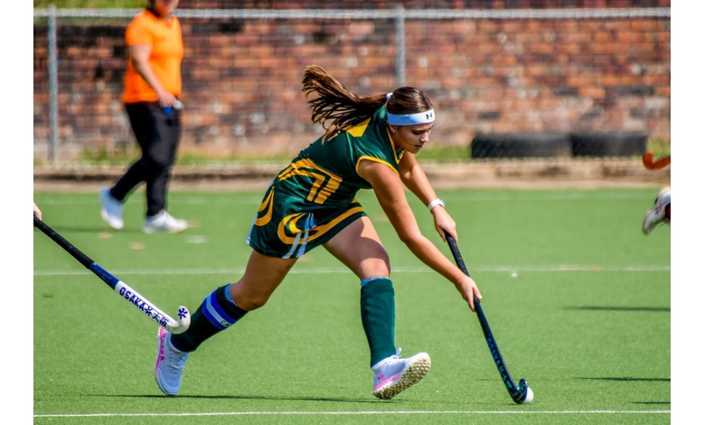 A female field hockey player in green and yellow uniform energetically dribbles a ball on a sunny field. An orange-clad referee watches in the background.
