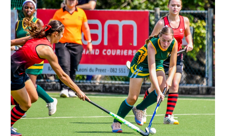 Two female field hockey players compete for the ball on a green field. One wears a red uniform, the other green. The background shows an orange-shirted referee and a red advertisement banner. The scene conveys action and intensity.