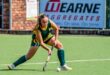 A female field hockey player in a green and yellow uniform focuses intently while striking a ball on a green field, with a brick wall in the background.