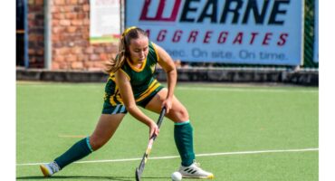 A female field hockey player in a green and yellow uniform focuses intently while striking a ball on a green field, with a brick wall in the background.