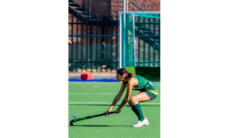 A female field hockey player in a green uniform poised to strike a ball on a green pitch. She is focused, with a goal visible in the background.