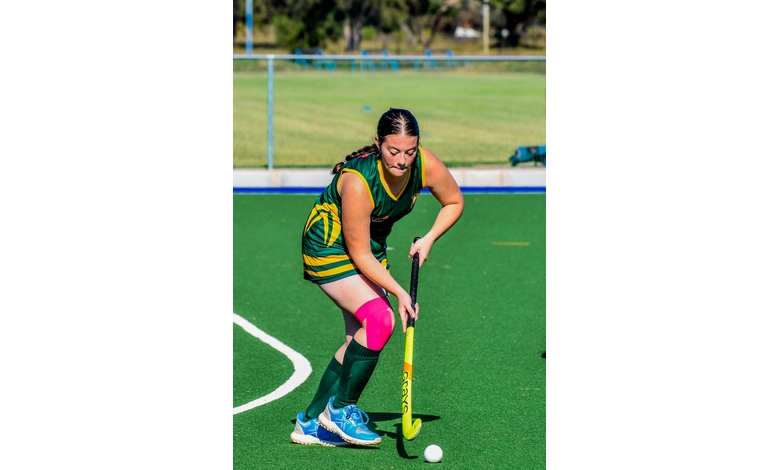 A female field hockey player in green-yellow gear is focused, controlling the ball with her stick on a sunny day. She wears a pink knee brace.
