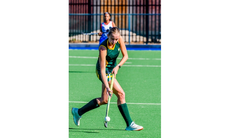 A field hockey player in a green and yellow uniform focuses intently as she maneuvers the ball with her stick. Another player is visible in the background.