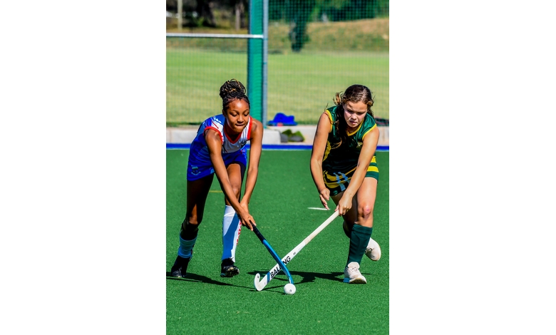 Two field hockey players compete for the ball on a green turf field. One in a blue uniform and the other in green, both focused and determined.