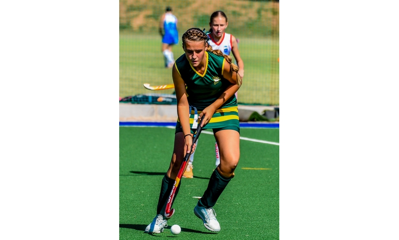 A female field hockey player in a green and yellow uniform focuses intently on controlling the ball with her stick during a match.