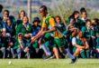 A field hockey player in green and yellow sprints with a stick during a match. A crowd of school students in uniforms watch and cheer energetically.