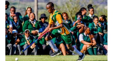 A field hockey player in green and yellow sprints with a stick during a match. A crowd of school students in uniforms watch and cheer energetically.