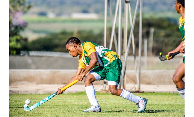 A field hockey player in a green and yellow uniform focuses intently, preparing to hit a ball with a blue stick on a grassy field under a bright sky.