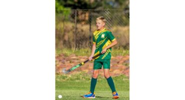A young boy in a green and yellow hockey uniform stands on a grassy field holding a hockey stick. He looks focused and ready to play.