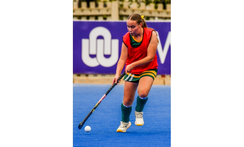 A female field hockey player, focused and determined, dribbles the ball on a blue turf. She's wearing a red jersey and green striped shorts.