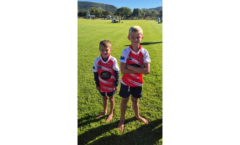 Two young boys in red and white sports jerseys stand on a grassy field, smiling at the camera. The sunny day enhances the cheerful atmosphere.