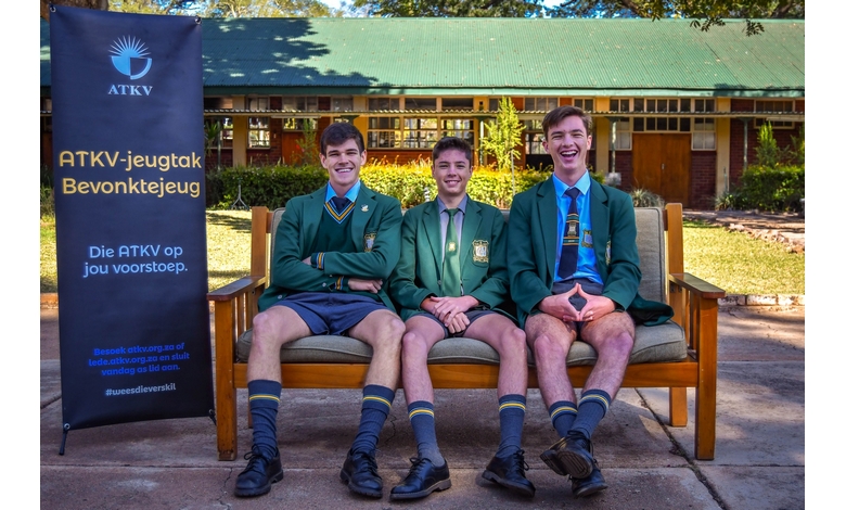 Three young men in school uniforms sit cheerfully on a wooden bench outdoors. A banner beside them reads "ATKV-jeugtak" against a school building backdrop.