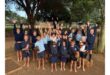 Children in school uniforms joyfully raise their arms while holding chickens. They stand barefoot outdoors, surrounded by trees and a fence, exuding excitement and happiness.
