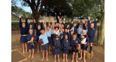 Children in school uniforms joyfully raise their arms while holding chickens. They stand barefoot outdoors, surrounded by trees and a fence, exuding excitement and happiness.