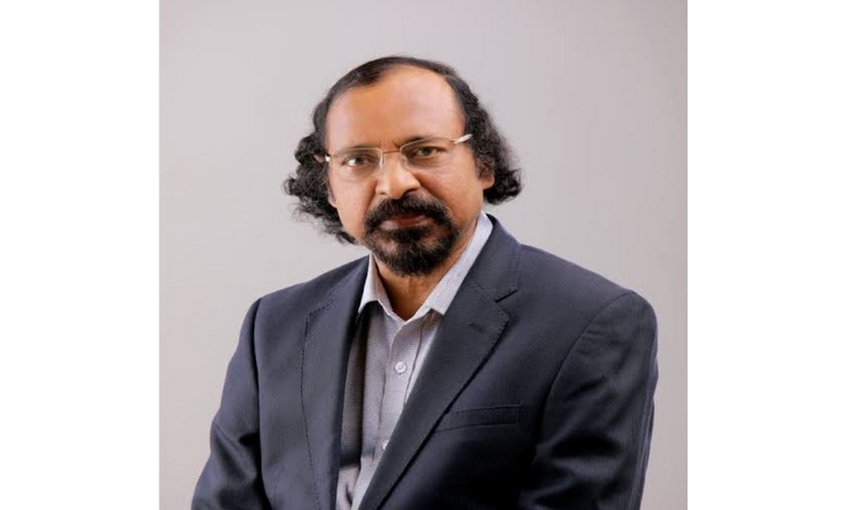Portrait of a man with glasses and curly hair, wearing a dark blazer and light blue shirt. He has a calm expression against a plain background.