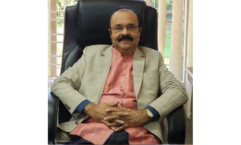 A person with a mustache wearing glasses sits comfortably in a black chair. They wear a cream blazer over a pink shirt, exuding a calm and confident demeanor.