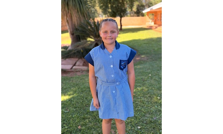Young girl stands smiling in a sunlit backyard, wearing a blue and white checkered school dress with a pocket. The mood is cheerful and relaxed.