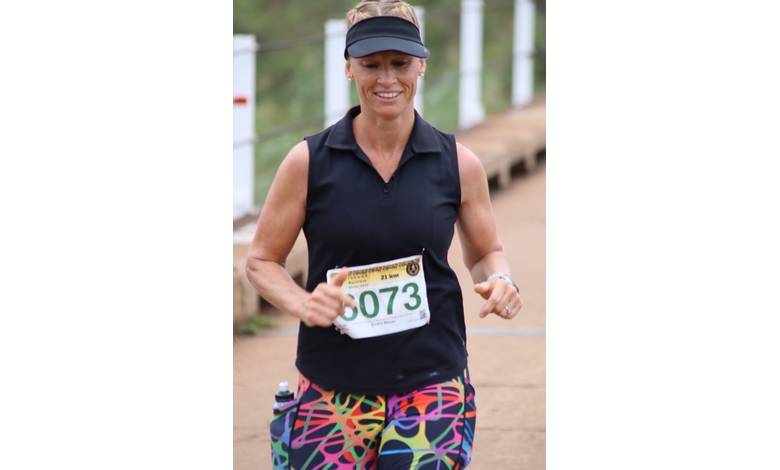 Smiling woman running on a path, wearing a black visor and top, colorful patterned shorts, and a race bib numbered 6073. Sense of determination.