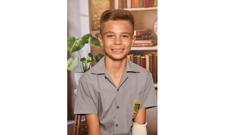 A smiling boy in a gray school uniform poses in front of a backdrop with books and plants. His left forearm is in a white cast. The atmosphere is cheerful and studious.