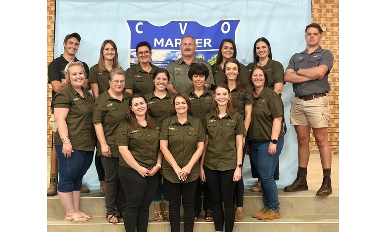 A group of 15 people wearing matching green shirts and posing in two rows. They're smiling in front of a banner that reads "CVO Marmer."