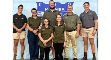 A group of seven smiling people in uniform stand in front of a blue banner. They wear green or grey shirts and shorts, conveying a unified and cheerful tone.