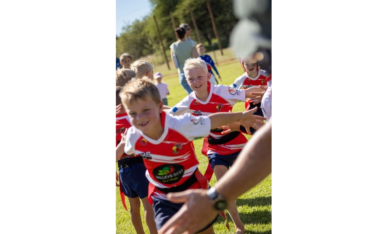Young boys in red sports jerseys run joyfully on a grassy field, smiling and reaching out for high-fives on a sunny day, conveying excitement and teamwork.