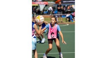 Young girl wearing a pink bib with "C" focuses intently on the netball, arms outstretched, on a sunny outdoor court. Background shows spectators.